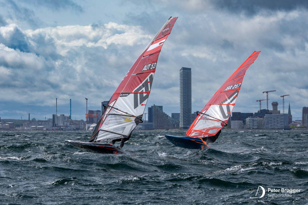 To vindsurfere sejler foran en bysilhuet under skiftende skyer.